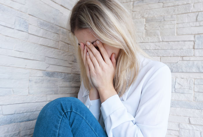 Woman sitting against a stone wall, covering her face with hands, showing regret and emotional distress.