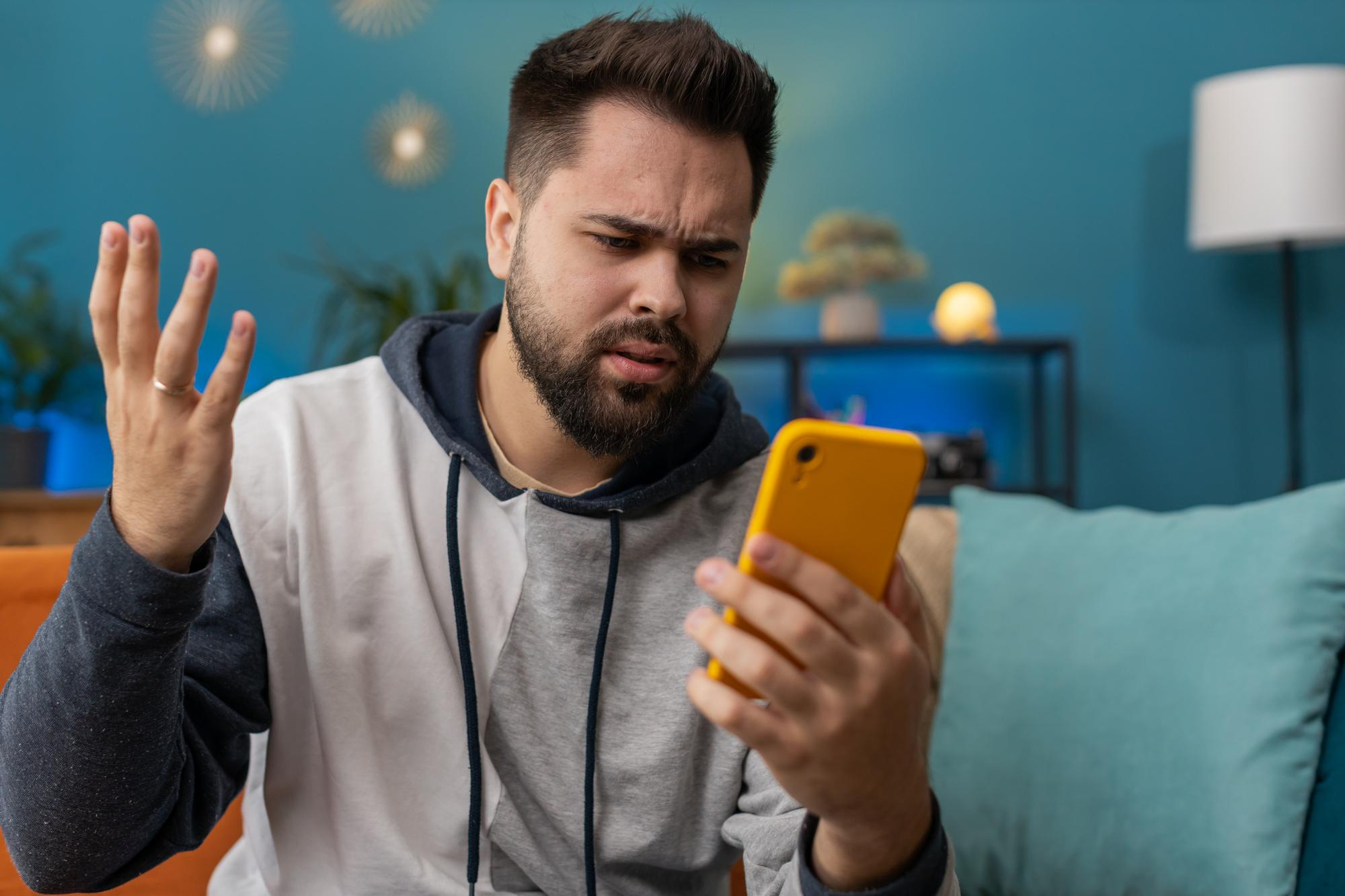 Frustrated man wearing hoodie looking at phone at home, reflecting stress of jobless conflict and family competition.