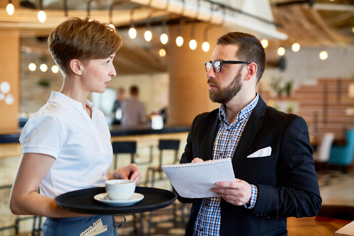 Man in glasses holding notes talking to waitress with coffee tray in a cafe, evoking haunted places chilling experiences.