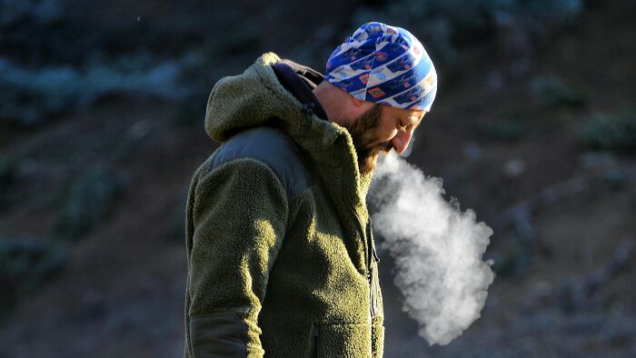 Man wearing a patterned beanie and green jacket outdoors, exhaling visible breath in cold air, childhood memories concept.