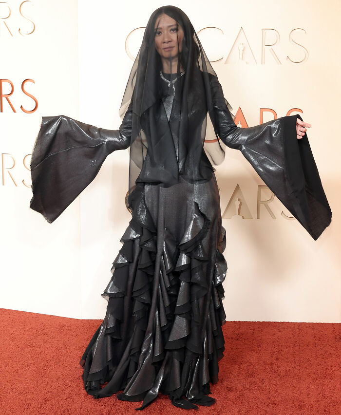 Woman on red carpet wearing a voluminous black ruffled gown with wide sleeves and a sheer black veil, fashion fails of award season.