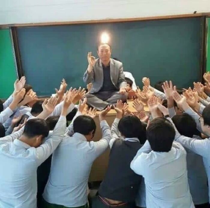 Man meditating on desk while group of people around him raise hands in creative poses indoors.