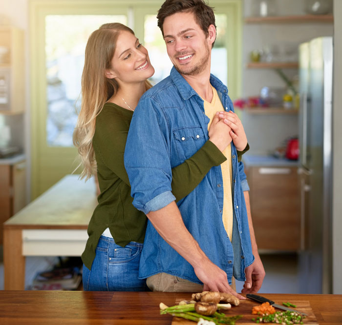 Couple embracing in kitchen while preparing food, illustrating woman trying to coexist with boyfriend&rsquo;s female friend.