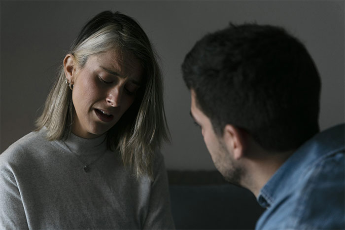 Woman admitting to cheating multiple times, looking upset while husband listens intensely in a dimly lit room.