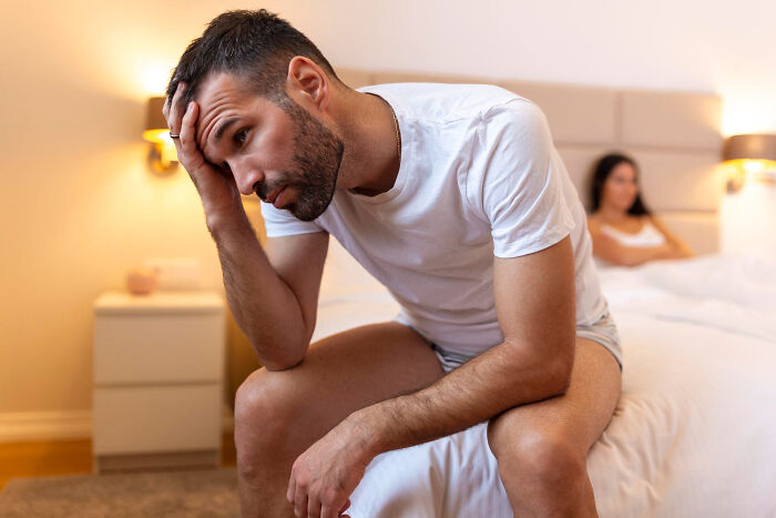 Man sitting on the edge of a bed looking stressed while a woman sits in the background, symbolizing women’s misunderstood experiences.