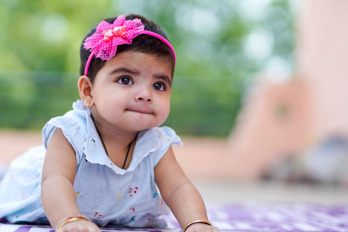 Toddler girl wearing a pink headband with a bow, looking curious while lying on her stomach outdoors in natural light.