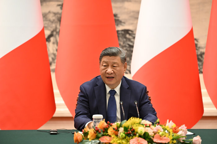 Xi Jinping speaking at a conference table with floral arrangement and large red and white flags in the background
