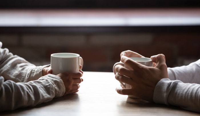 Two women holding white mugs, sitting across a wooden table in a dimly lit, tense conversation setting.