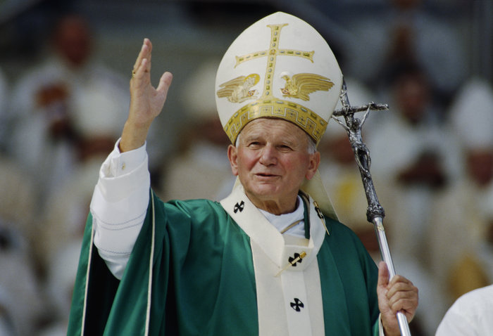 John Paul II in green and white religious robes raising hand, holding ceremonial staff during public event.