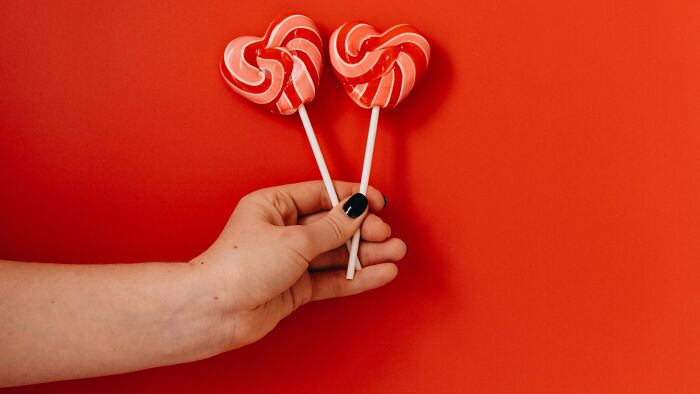 Hand holding two heart-shaped red and white lollipops against a solid red background, childhood memories concept.