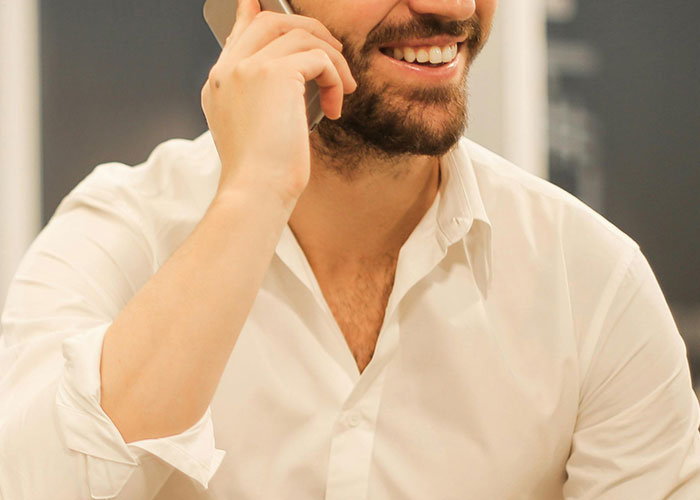 Bearded man in white shirt smiling while talking on a phone, hinting at a messy love triangle situation.