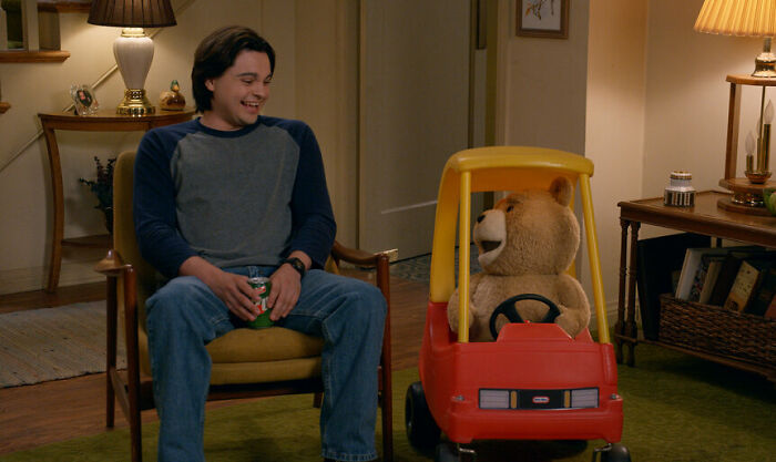 Man sitting on a chair smiling at a talking teddy bear in a toy car, related to Seth Macfarlane Ted Season 2 news.