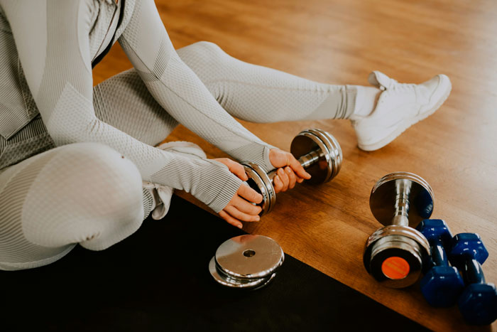 Woman in light workout clothes sitting on floor holding dumbbell with more weights around, focusing on fitness routine Woman in light workout clothes sitting on floor holding dumbbell with more weights around, focusing on fitness routine