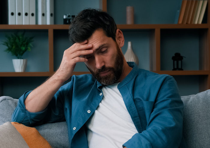 Homem de camisa azul parecendo estressado em um sofá, refletindo sobre como impedir os pais de serem babás depois de ouvirem seus pensamentos Homem de camisa azul parecendo estressado em um sofá, refletindo sobre como impedir os pais de serem babás depois de ouvirem seus pensamentos