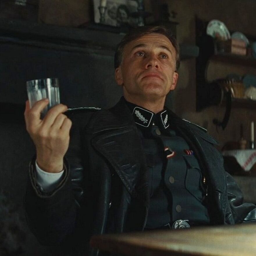 Actor in a supporting performance, dressed in a dark military coat, holding a glass and looking thoughtfully upward.