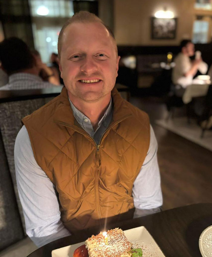 Man smiling at a restaurant table with dessert, representing family's shocking decision after students' prank on teacher tragedy.