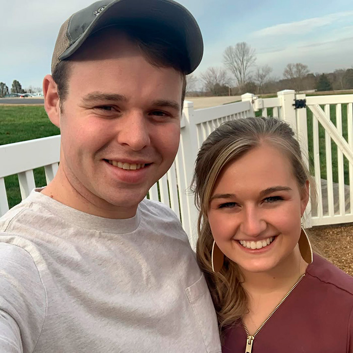 Young couple smiling outdoors by white fence with green fields in the background related to Kendra Duggar mugshot news.