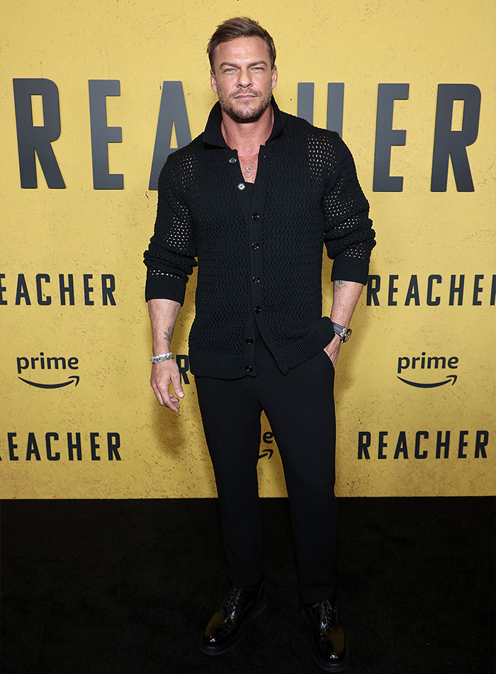 Alan Ritchson posing at a Reacher event, wearing a black buttoned sweater and black pants against a yellow backdrop.
