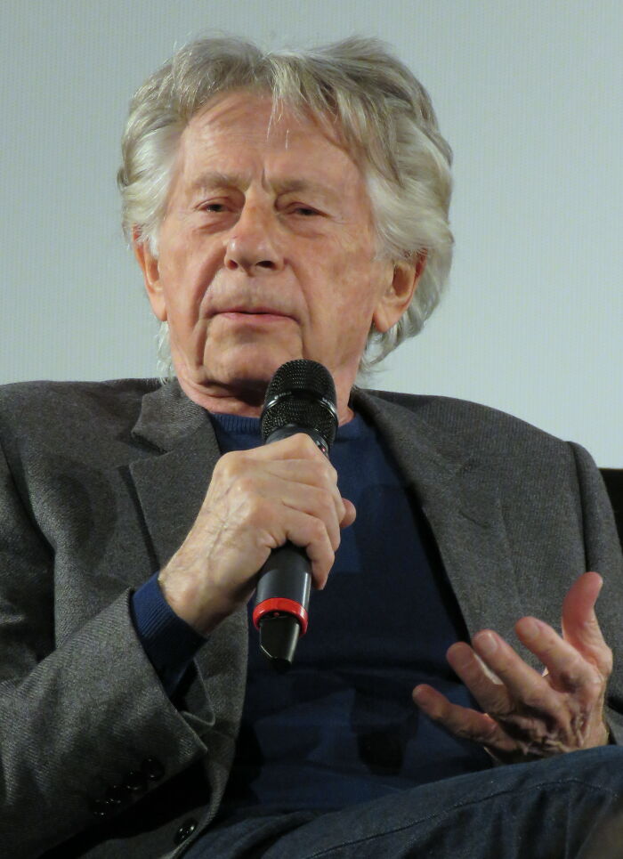 Older male celebrity speaking into a microphone during an event, related to celebrities people used to like but now hate.