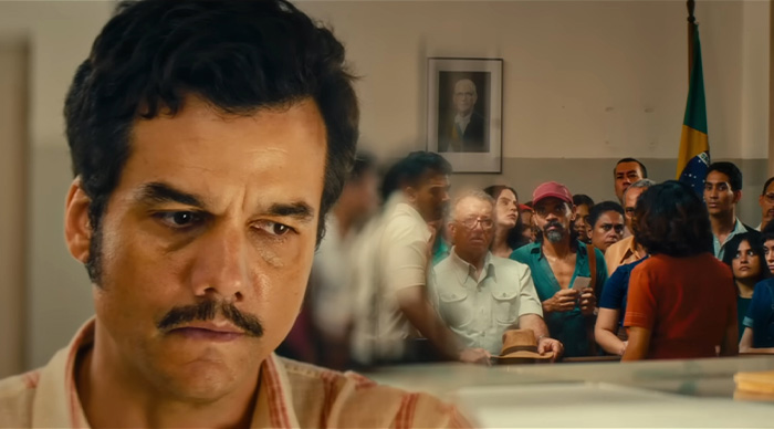 Man with mustache in striped shirt looking down thoughtfully while a crowd gathers behind him indoors, Oscars prediction theme.