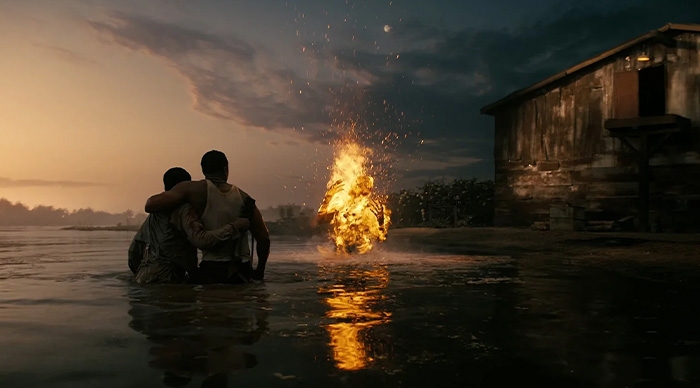 Two people in water at dusk watching a figure engulfed in flames near a rustic cabin for Oscar predictions 2026.
