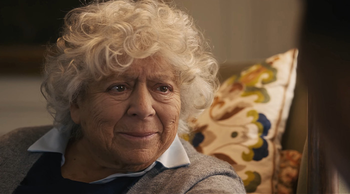 Elderly woman with curly white hair smiling gently during a relaxed conversation about Oscar predictions and community trends.