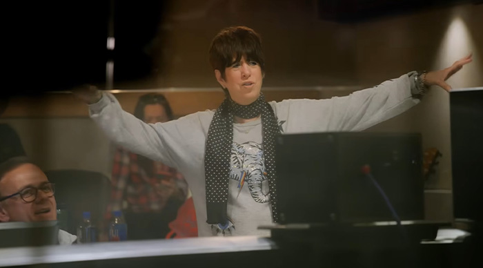 Person wearing a gray sweatshirt and polka dot scarf gesturing with arms in a studio for Oscar predictions and community trends.