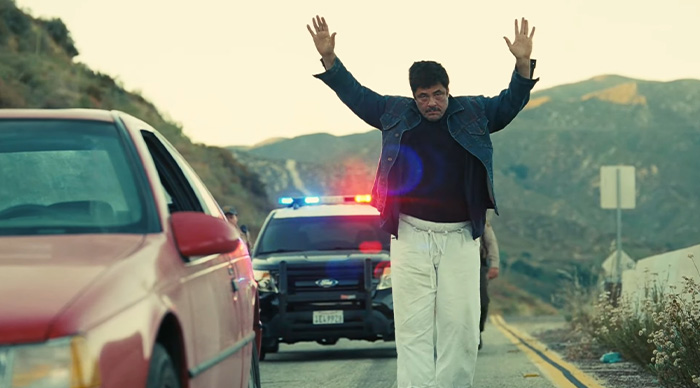 Man with raised hands near red car, police car with flashing lights behind, scenic mountainous road, 2026 Oscar predictions scene.