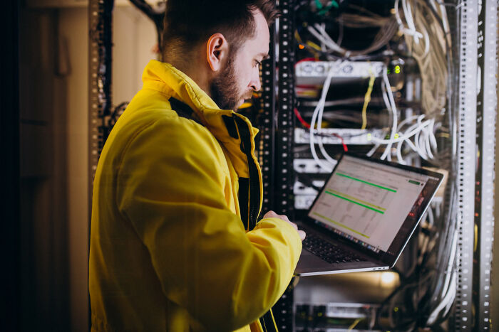 Man in yellow jacket checking laptop near server equipment in a dimly lit basement with network cables and connections.
