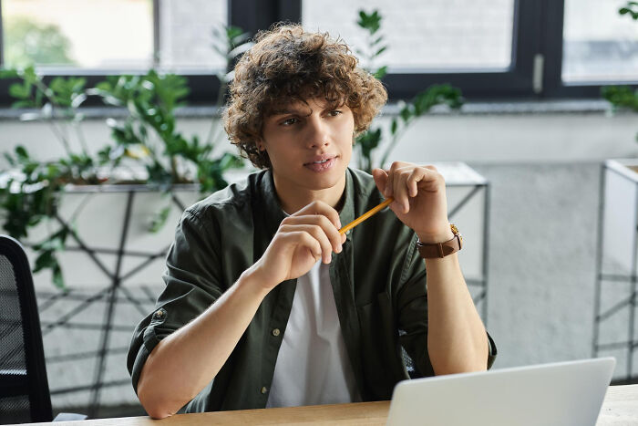 Young man with curly hair holding a pencil, seated indoors, reacting with a thoughtful expression to Gen Z stare discussion.