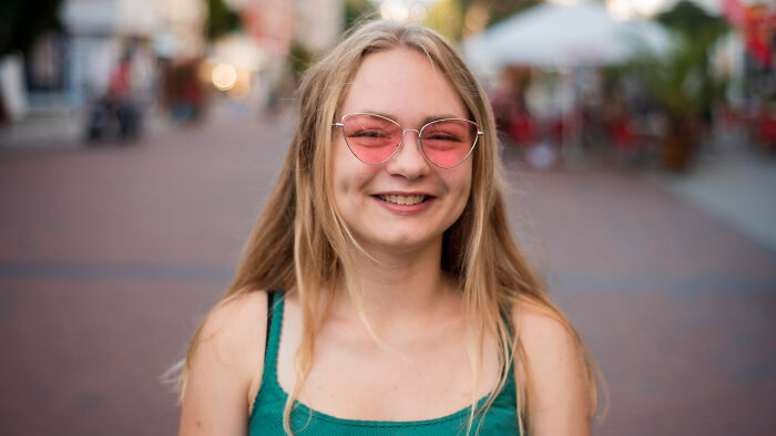 Young woman with long blonde hair wearing pink sunglasses and a green top smiling outdoors, reflecting Gen Z stare reactions.