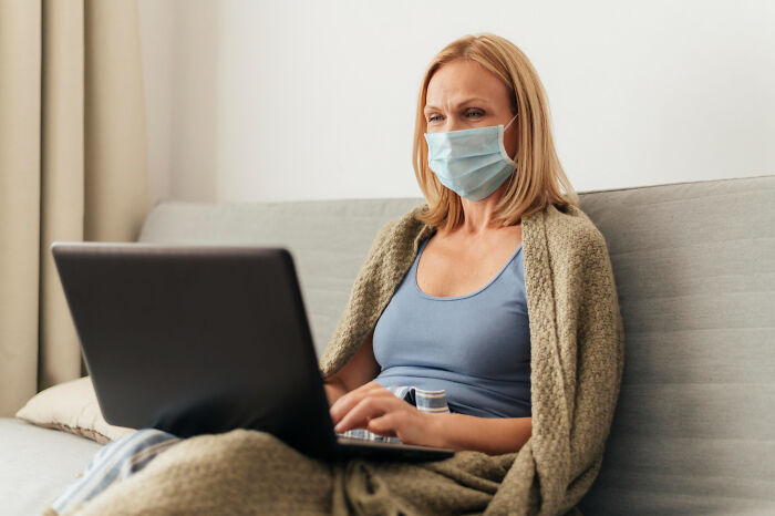 Woman wearing mask and wrapped in blanket using laptop, illustrating doctors hiding truths about lost lab samples from patients.
