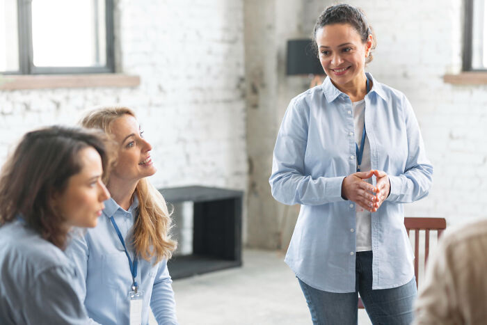 Three women in casual workwear discussing shaky industries with one woman standing and smiling confidently in a bright office.