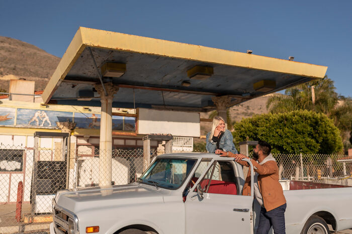 Young woman and man sharing a moment by a vintage truck near an abandoned gas station, a hilarious culture shock scene outdoors.