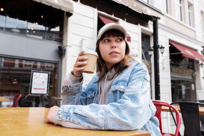 Young woman in a denim jacket holding a coffee cup, sitting outdoors and showing a thoughtful Gen Z stare.