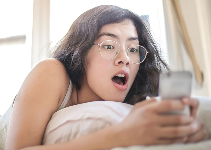 Young woman with glasses lying on bed, looking shocked at her phone, reflecting chaotic secrets of a messy love triangle.