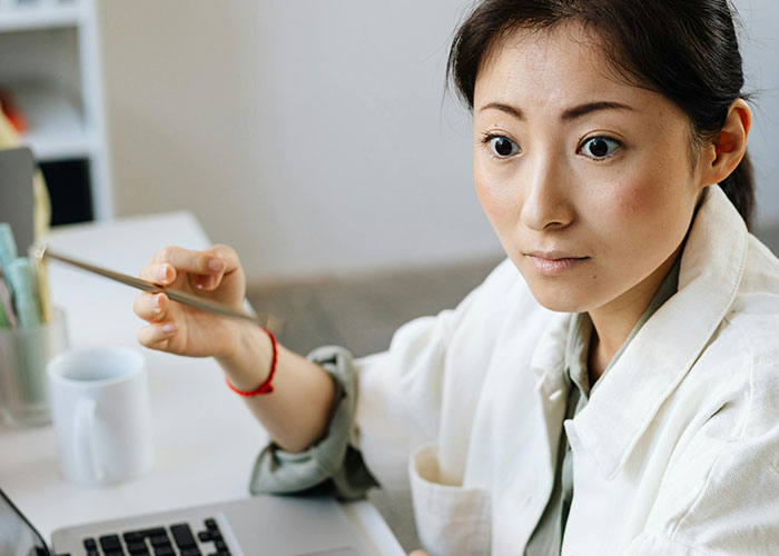 Young woman sitting by a laptop, holding a pencil, deep in thought about a messy love triangle and chaotic secrets.