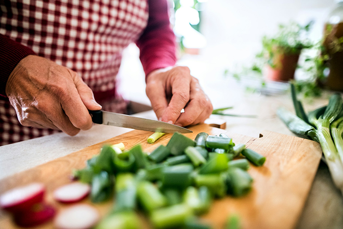 Person cutting green vegetables on a wooden board, showing hands slicing fresh produce in a bright kitchen.