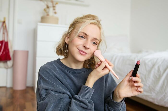 Woman applying makeup with brush and lipstick in a cozy bedroom, illustrating secrets women don’t tell men.