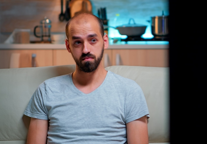 Man with one arm sitting on a couch looking frustrated in a kitchen setting, illustrating challenges with cutting vegetables.