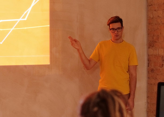 Young man in a yellow shirt presenting a messy love triangle concept with chaotic secrets on a projected screen during a meeting.