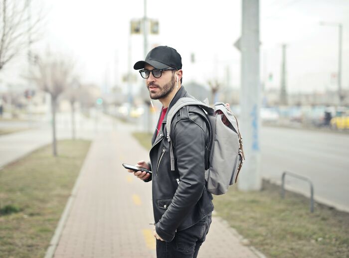 Man wearing glasses and a black cap standing on a sidewalk, holding phone and wearing a gray backpack in an urban setting.