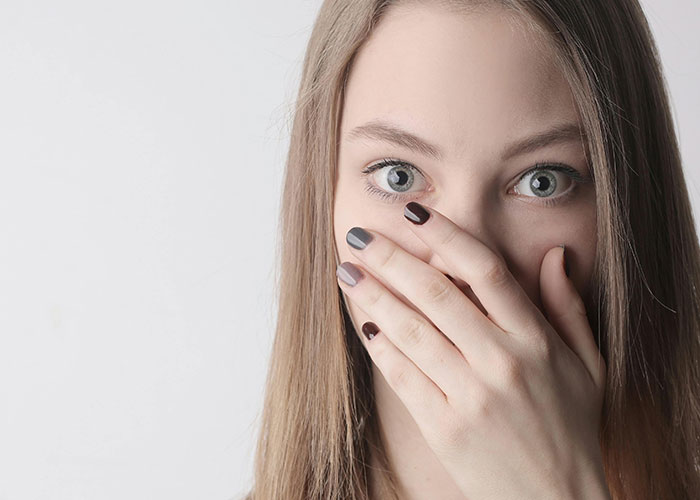 Young woman with multicolored nails covering her mouth, expressing surprise related to a messy love triangle secret.