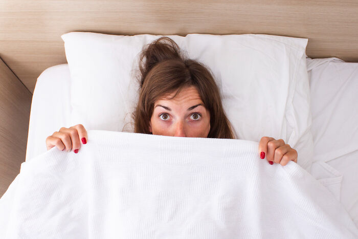 Woman hiding under white blanket in bed with wide eyes, illustrating spine-chilling experiences in haunted places.