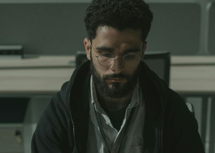 Man with glasses and beard sitting indoors, portraying deep thought about love triangle secrets and chaotic relationships.