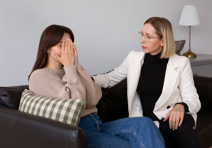 Two women having a tense conversation on a couch, one covering her face, showing conflict among friends on vacation plans.