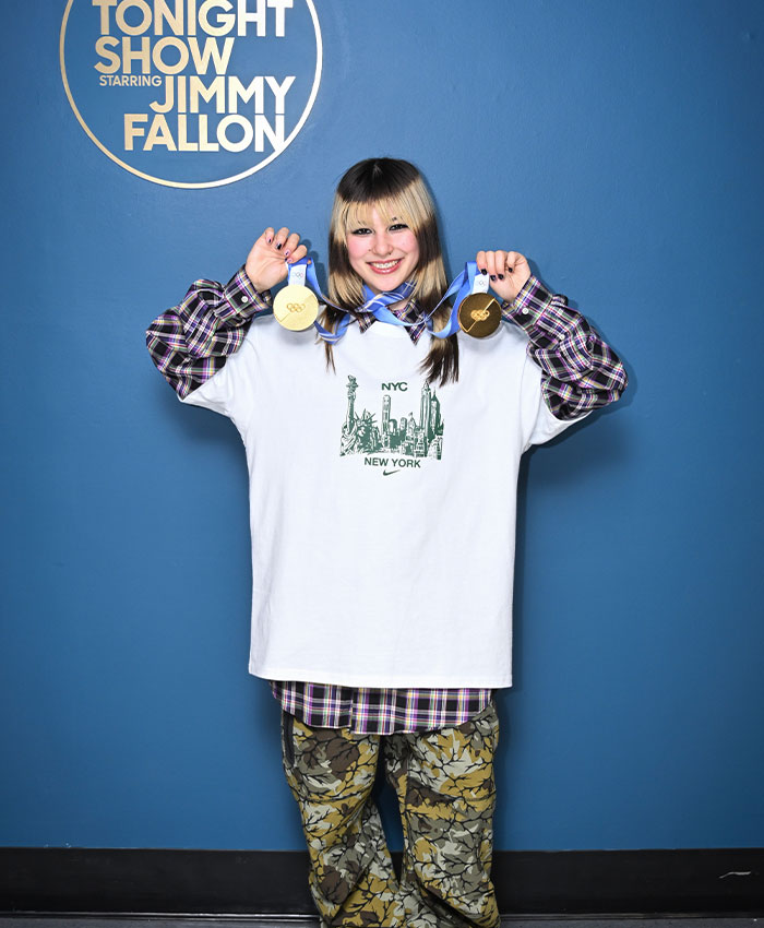 Olympic hero Alysa Liu smiling and holding two gold medals while standing against a blue Tonight Show backdrop.