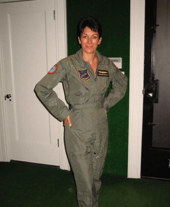Ghislaine Maxwell standing in a military-style flight suit, posing indoors with hands on hips, smiling confidently.