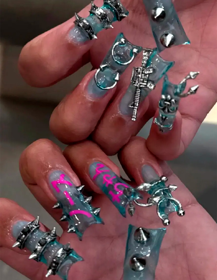Close-up of spiked and pierced manicure with silver embellishments and neon pink writing on long clear nails.