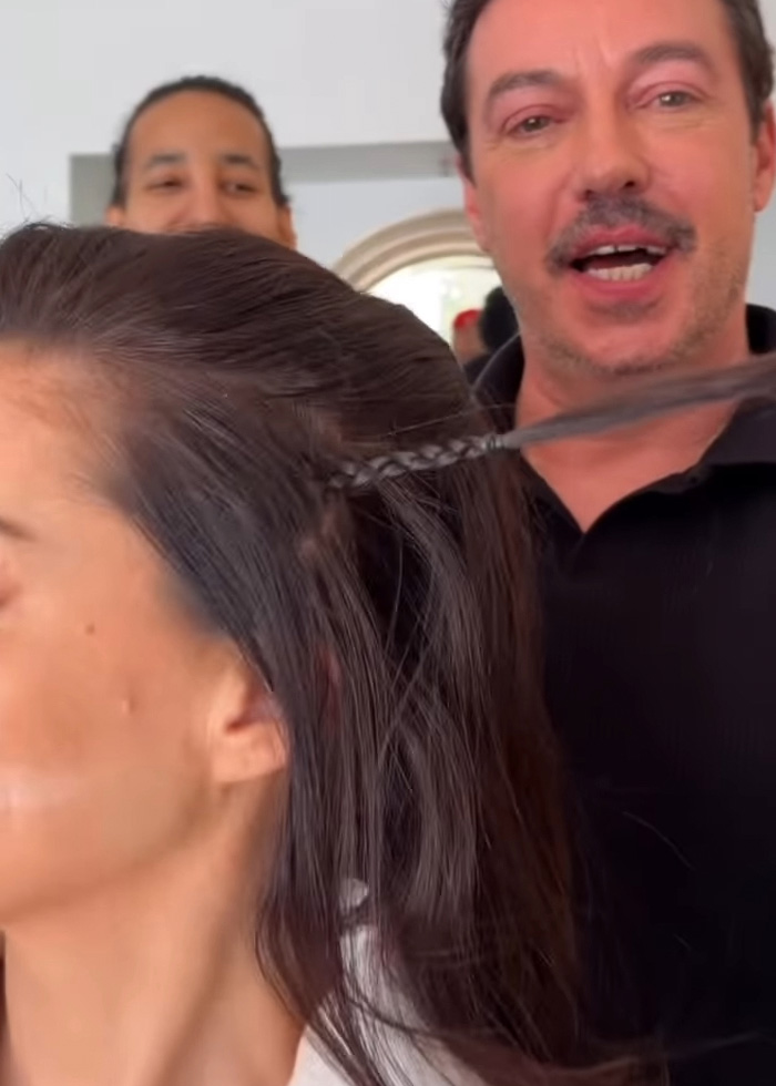 Cabeleireiro demonstrando um truque de cabelo no cabelo escuro de uma mulher, relacionado à revelação do rosto tenso de Anne Hathaway.